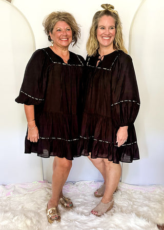 knee length black tiered dress with smocking on the chest and a tie closure at the rounded neck. 3/4 sleeve, leopard piping.