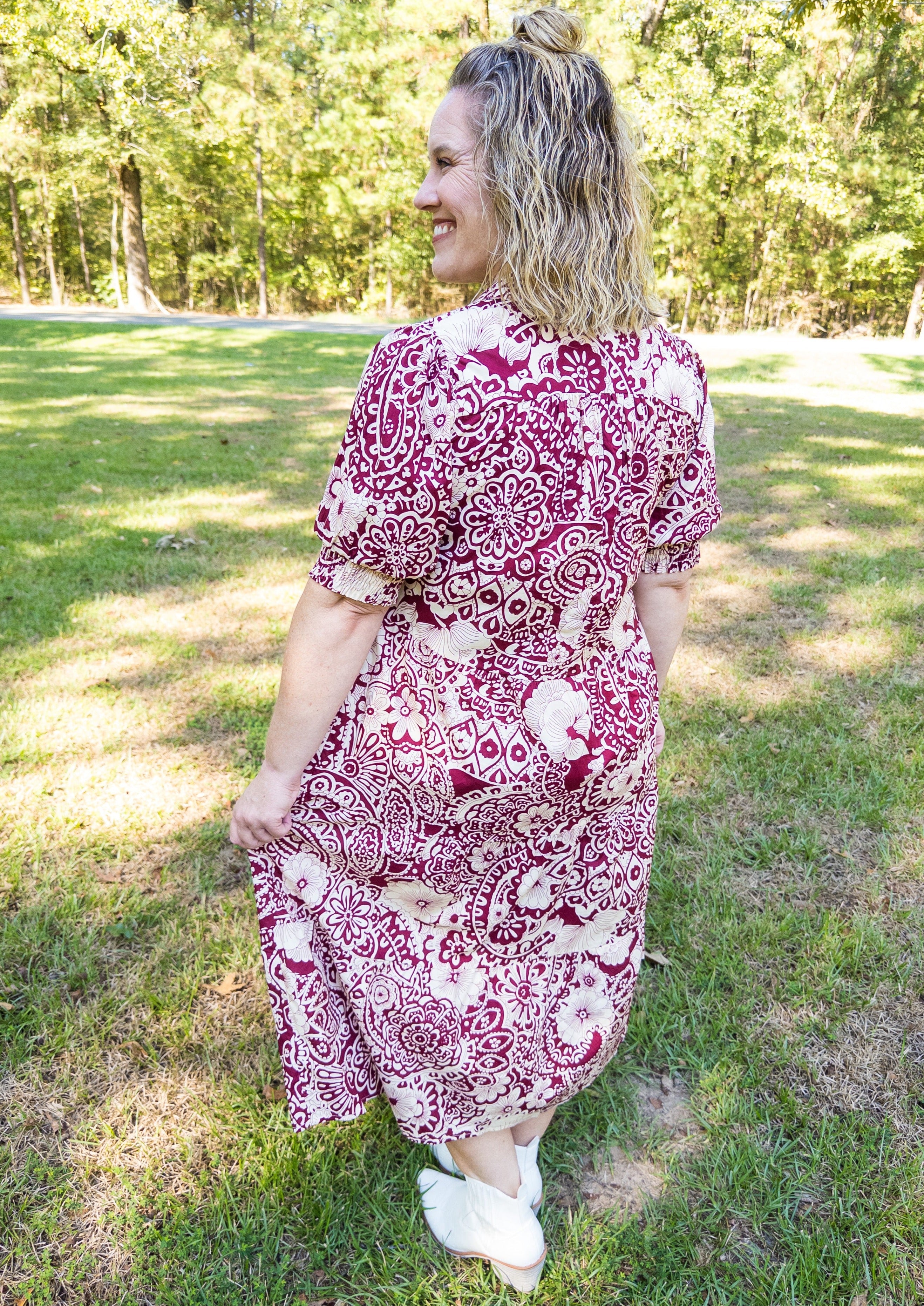 Back view of off white abstract design with flowers on a sangria/wine colored background. Midi length, below the knee. V-neckline with collar. Little balloon sleeve with wide elastic banded cuff above elbow.