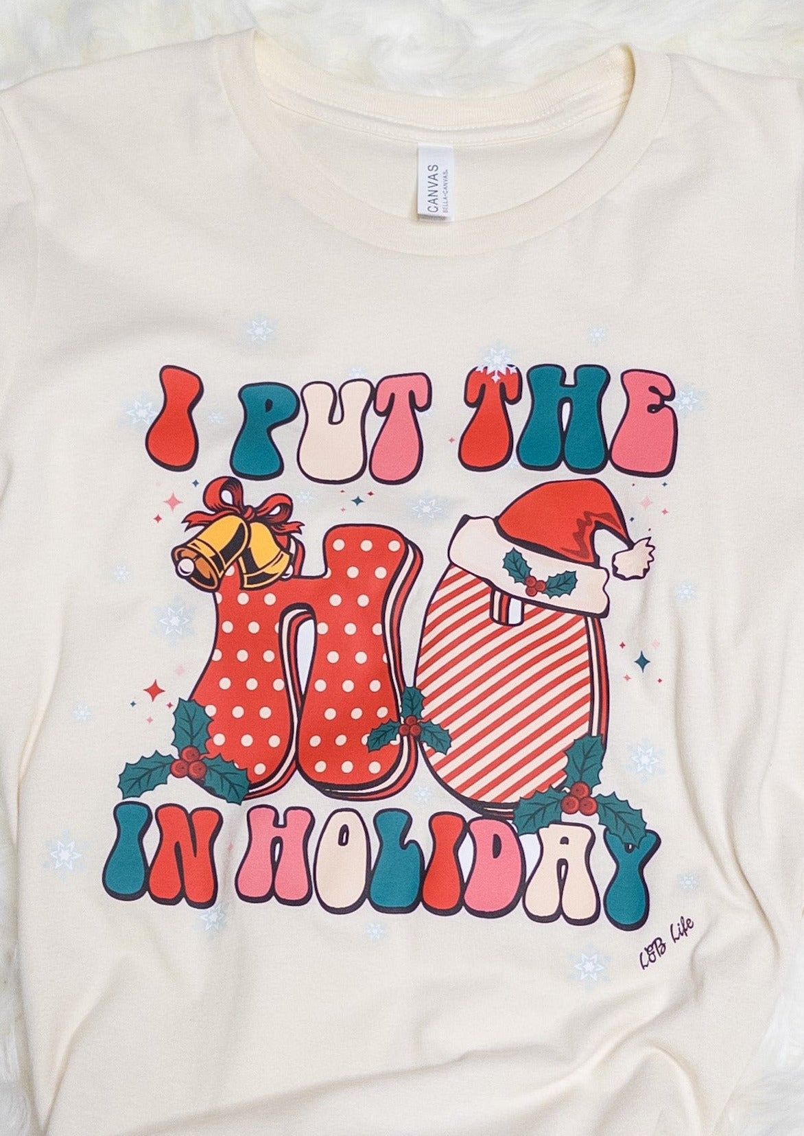 Cream colored tee shirt that says "I put the ho in holiday". Festive red and green bubble writing, boughs of holly, a santa hat, and 2 bells tied with ribbon.