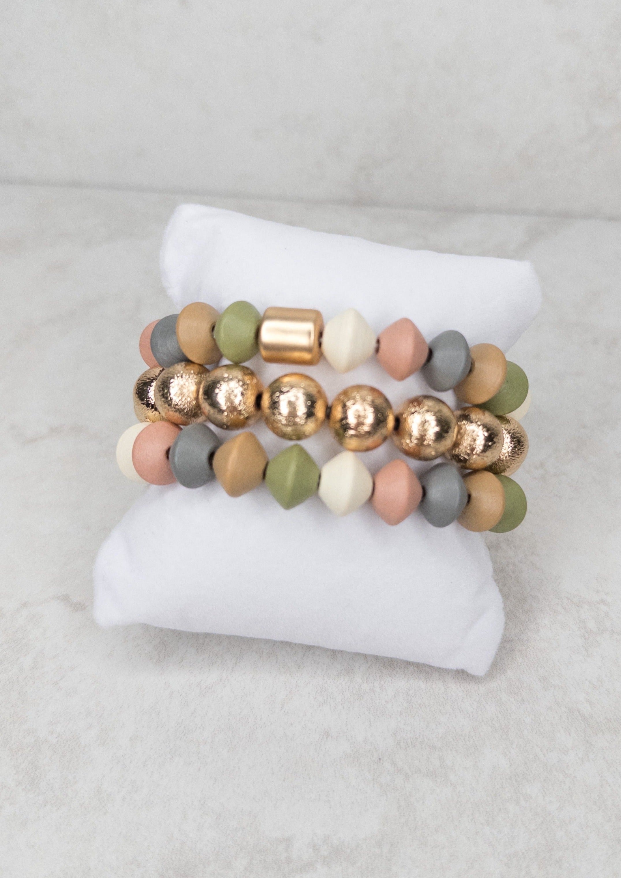 3 bracelets on a bracelet pillow. Top and bottom bracelet have pastel colored beads. The middle bracelet has round, gold beads.