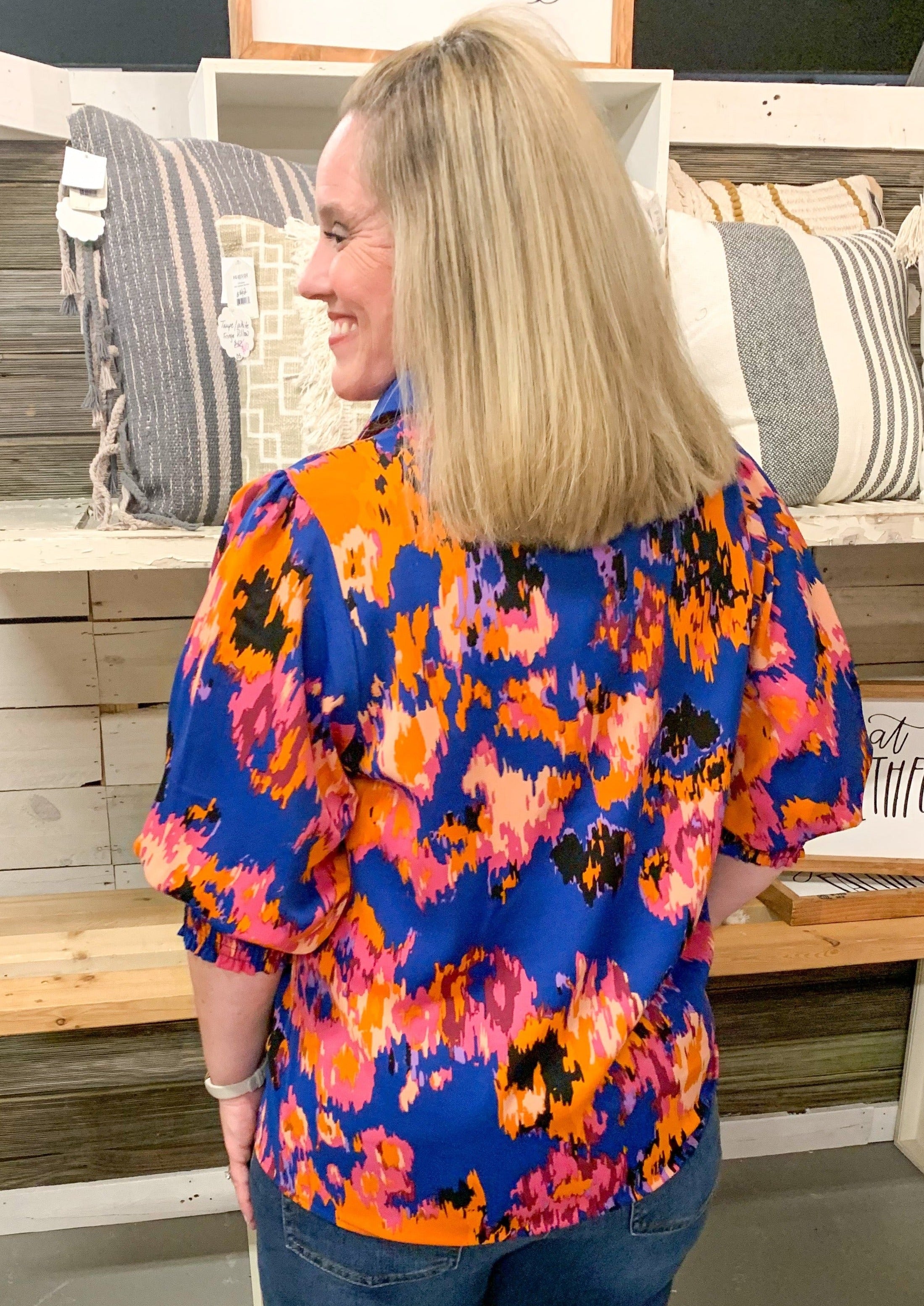 back of blue pink yellow and orange abstract print satin blouse. Has slight V neck, collar, and a puff sleeve with a wide elastic banded cuff near elbow.