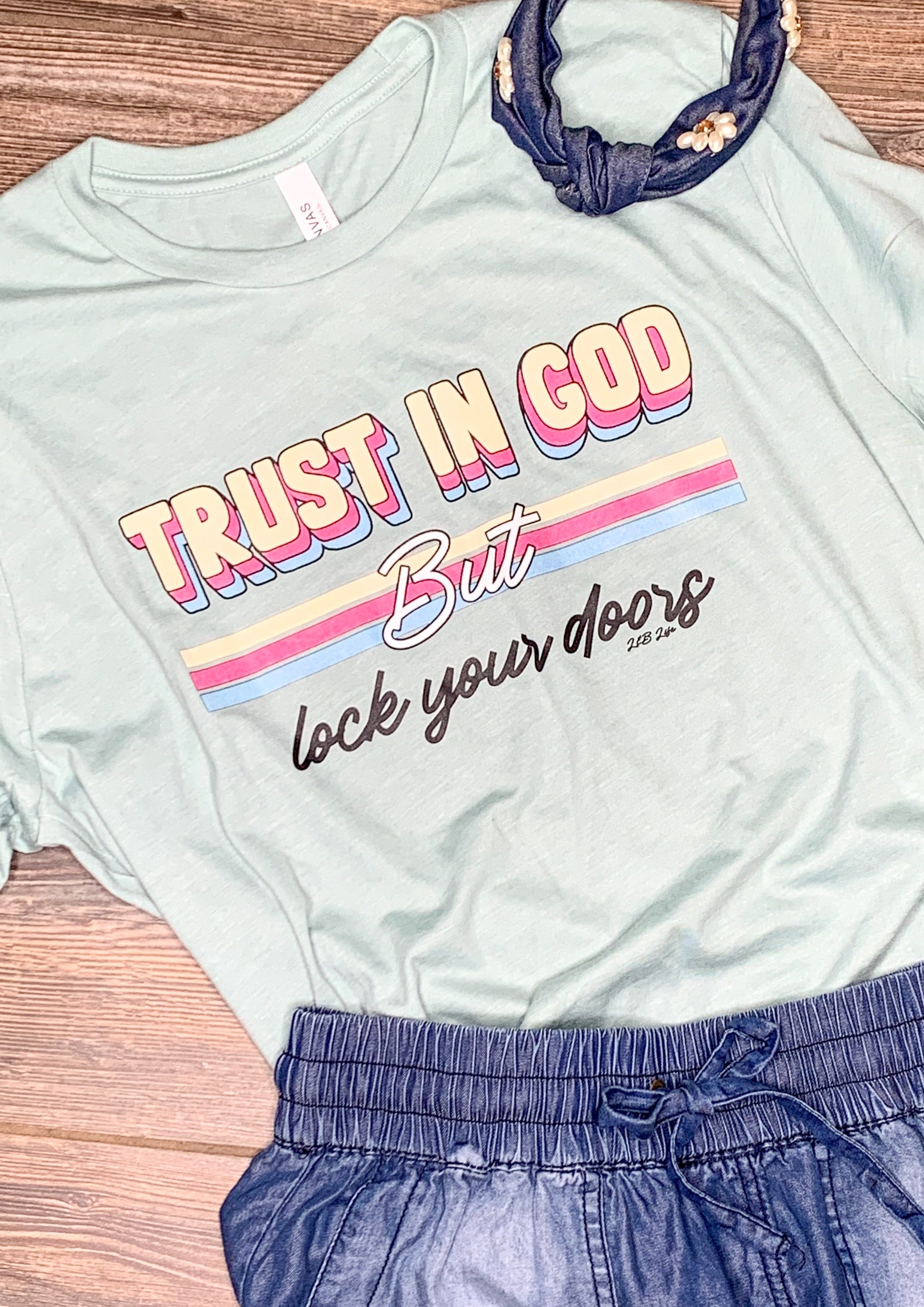 A light dusty blue tee shirt that says "Trust in God but lock your doors" with the colors black, white, pink, blue, and yellow.