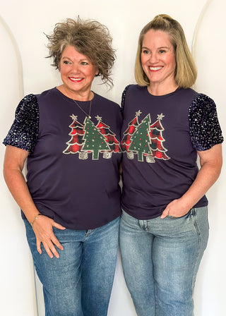 Two women wearing a navy short sequin sleeve top with plaid and green embellished trees