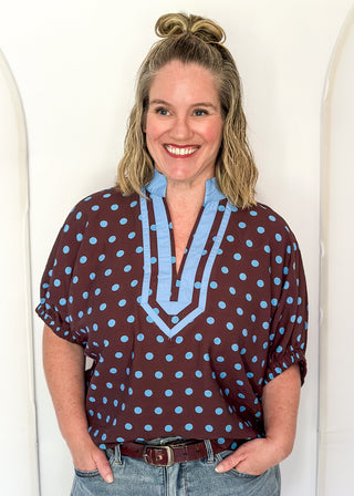 A woman smiling and wearing a brown short-sleeve blouse with light blue polka dots and a light blue V-neck trim, paired with blue jeans, standing against a white arched wall.