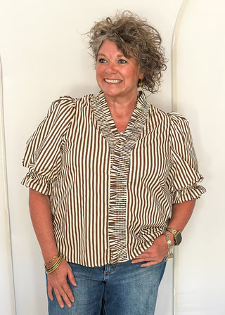 A smiling woman with curly gray hair wearing a brown and white vertically striped blouse with ruffles and puff sleeves, paired with blue jeans, standing against a white arched backdrop.