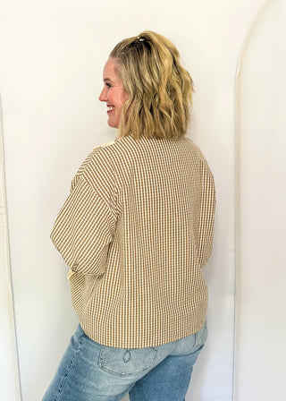 A woman with blonde hair tied back is shown from the back wearing a beige and white gingham patterned long-sleeve top and blue jeans, standing against a white studio backdrop with arch-shaped panels.