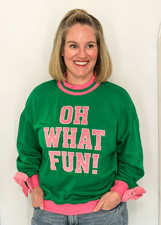 woman wearing a green long sleeve mock neck top with pink contrast sleeve  and "OH WHAT FUN" embroidered letter patches.