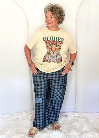 A woman modeling a cream graphic t-shirt with a leopard print and the word 'BOUIFE' in green, paired with blue and black plaid pants, standing against a white arched backdrop.