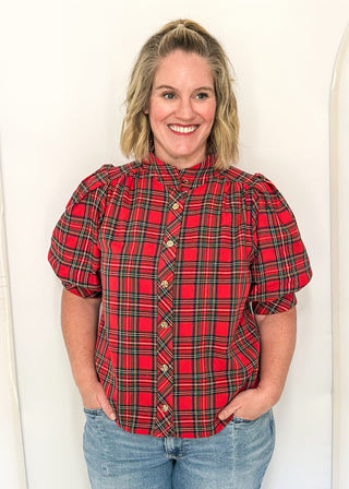 A smiling woman with blonde hair wearing a red and green plaid puff-sleeve blouse with gold buttons, paired with blue jeans, standing against a white arched backdrop.
