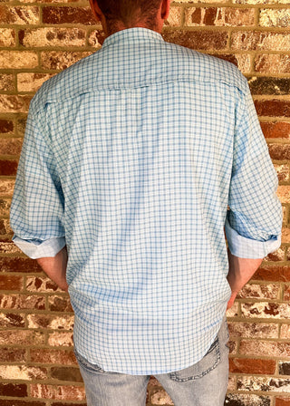 A man viewed from behind wearing a light blue and white gingham long-sleeve button-down shirt, light-wash jeans, and a cream cowboy hat, standing against a rustic brick wall.