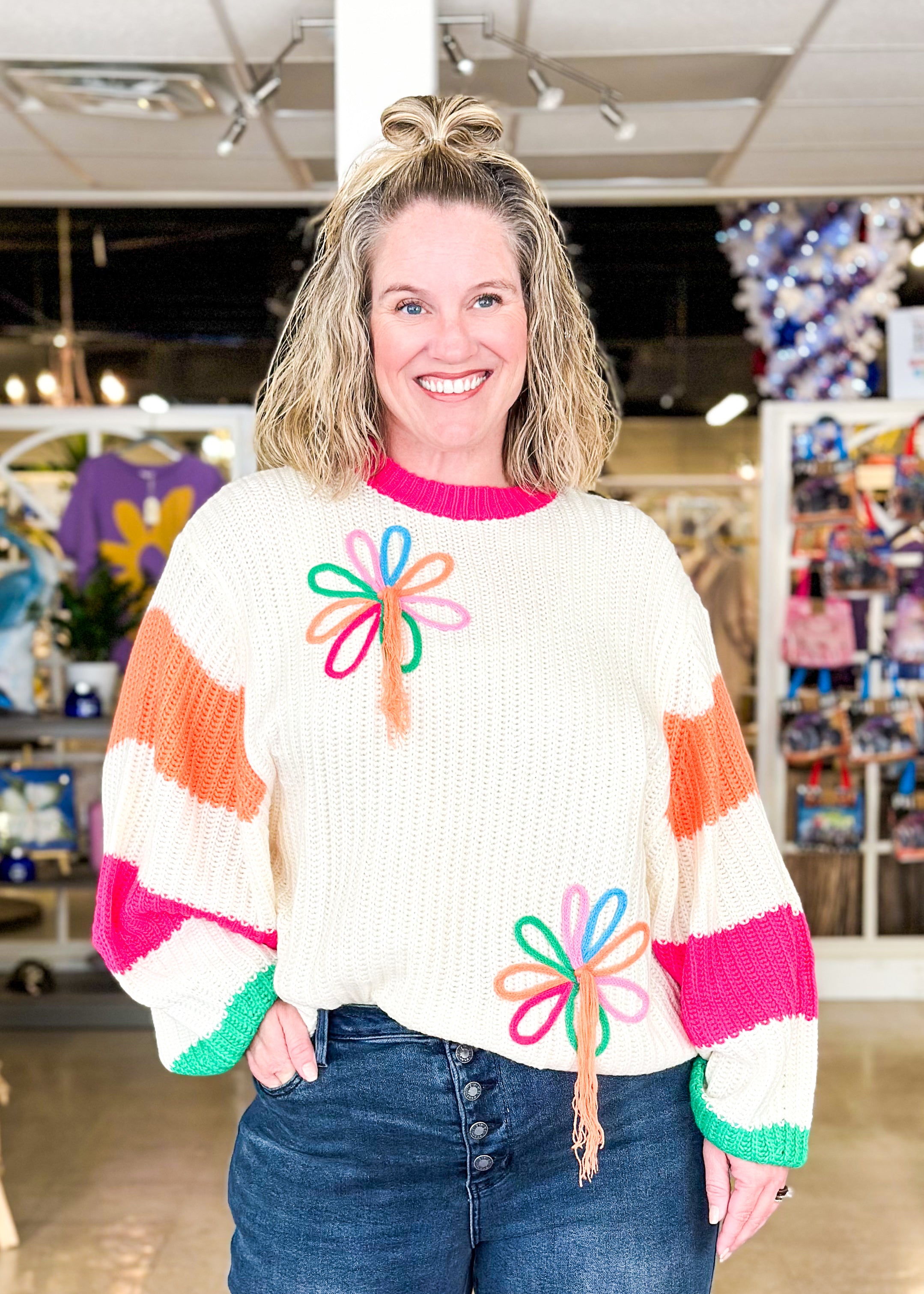 cream sweater with 2 bright flowers and orange tassels hanging from them, sleeves have wide orange and pink stripe, green around cuff and pink around bottom and neck