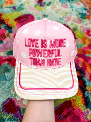 A pink snapback cap with white polka dots on the front panel and a cream-and-white chevron patterned visor, held up by a hand. The front features the embroidered text 'LOVE IS MORE POWERFUL THAN HATE' in pink.