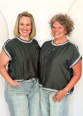Two women standing side-by-side wearing matching dark gray short-sleeve tops with white and blue striped trim, paired with light-wash blue jeans, against a white studio backdrop.