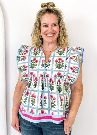 Flutter sleeve top with off white background and soft blue grid pattern-large magenta florals with green stems/leaves in each grid.  Soft blue ribbon trim on sleeve and collar.  Soft blue ribbon tie neck.  Soft blue and magenta trim on bottom band. 