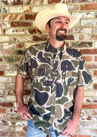 A man wearing a beige cowboy hat and a short-sleeve button-up shirt with a green, brown, and black camouflage pattern, standing against a rustic brick wall.