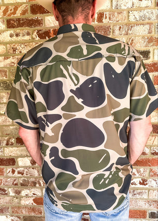 A man viewed from behind wearing a short-sleeve button-up shirt with a bold organic camouflage pattern in beige, olive green, and dark charcoal gray, paired with blue jeans against a brick wall background.