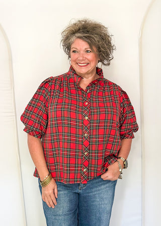 A woman with short, curly gray hair stands smiling, wearing a red and black plaid short-sleeve button-up shirt with blue jeans, posed against a white studio backdrop with arched panels.