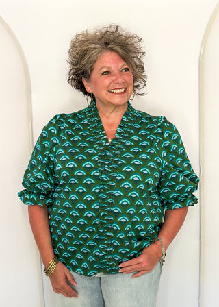 A smiling woman with short curly gray hair wearing a green short-sleeve blouse with a teal and black scallop pattern, paired with light blue jeans, standing against a white arched backdrop.