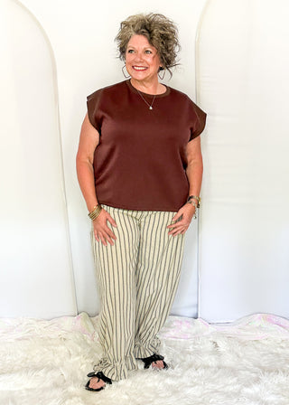 A woman modeling a brown short-sleeve satin top and beige-and-black striped pants, standing against a white arched backdrop on a white faux fur rug.