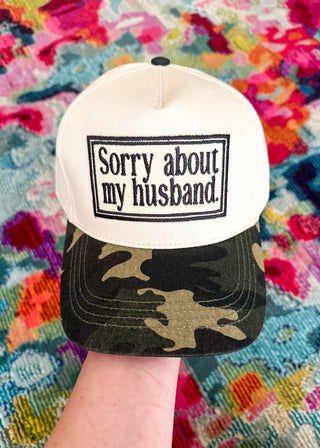 A white snapback cap with a black embroidered patch reading 'Sorry about my husband.' on the front, paired with a dark camouflage-patterned brim, held by a hand against a colorful patterned textile background.