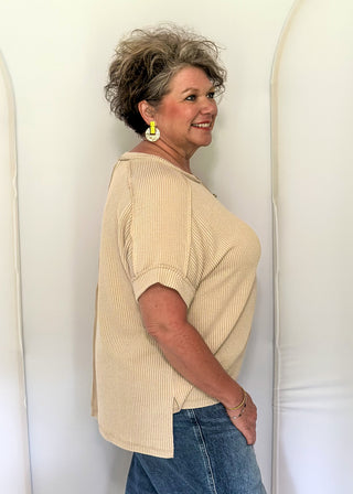 Woman wearing a beige sweater and blue jeans standing against a white wall.