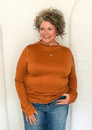 A smiling woman with curly gray hair stands against a white arched backdrop, wearing a rust-orange long-sleeve knit top and blue jeans.