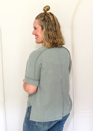 Person wearing a green checkered shirt and blue jeans standing against a white wall.
