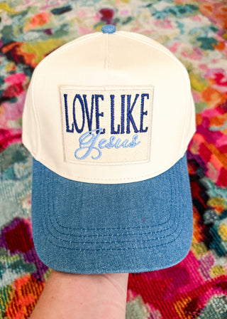 A two-tone baseball cap held by a hand, featuring an off-white crown with a rectangular patch that reads 'LOVE LIKE Jesus' in navy and light blue embroidery, and a blue denim-style curved brim. The background is a colorful, textured blanket.