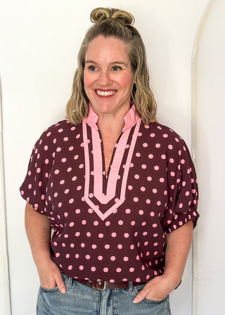 A smiling woman with blonde hair in a bun wears a short-sleeve blouse with pink polka dots and a pink chevron collar detail on a burgundy base, paired with blue jeans, standing against a white arched backdrop.