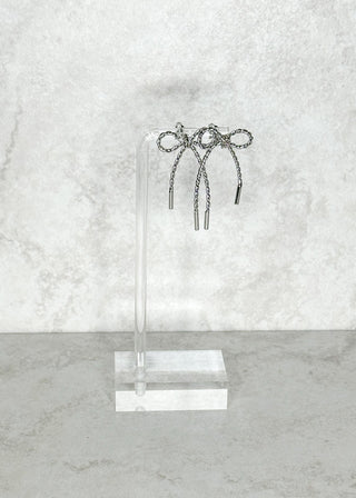 Silver bow Earrings on a clear stand against a light gray marble background
