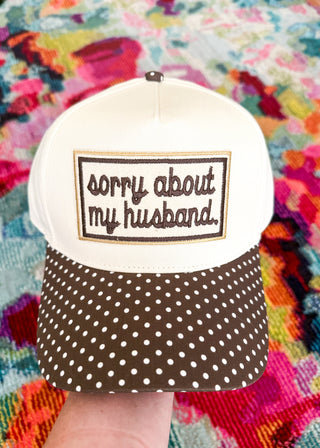 A white and brown baseball cap held by a hand, featuring a white front panel with the embroidered text 'sorry about my husband.' in brown script inside a brown border, and a brown brim covered in white polka dots, set against a colorful, textured background.