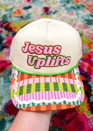 A cream-colored baseball cap with 'Jesus Uplifts' embroidered on the front, held by a hand against a vibrant, multicolored patterned background.