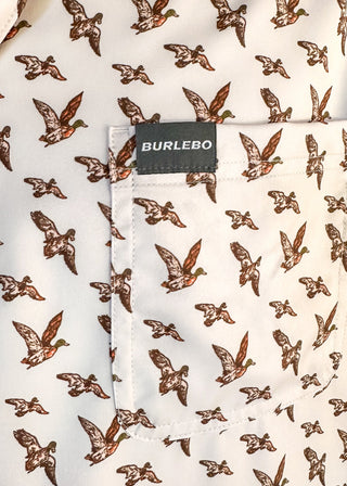 Close-up of a white short-sleeve button-up shirt with an all-over brown bird print and a chest pocket. A black BURLEBO brand label is stitched onto the pocket.