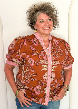 woman standing in a rust, pink and off white button up top and jeans.
