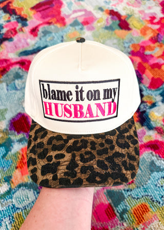 A white baseball cap with a leopard-print brim and a front patch reading 'blame it on my HUSBAND' in black and pink text, held by a hand against a colorful patterned fabric background.