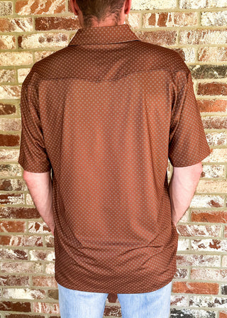 A man viewed from behind wearing a brown short-sleeve button-up shirt with a small white geometric pattern, light blue jeans, and a beige cowboy hat, standing in front of a brick wall.