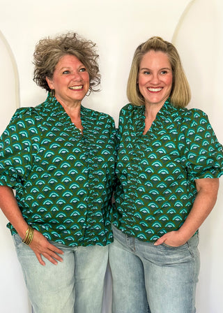 Two women standing side by side wearing matching green short-sleeve button-up shirts with a repeating teal and green scallop pattern, paired with light-wash jeans, against a white arched backdrop.
