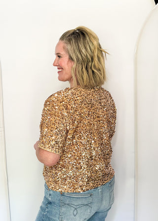 A woman with blonde hair is shown from the back wearing a short-sleeve top covered in gold sequins, paired with light blue jeans, standing against a white studio backdrop.