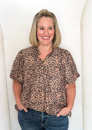 A smiling blonde woman wearing a brown and black leopard print short-sleeve blouse with blue jeans, standing against a white arched studio backdrop.