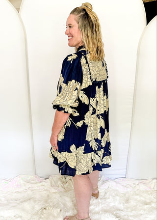 Woman wearing a navy floral dress standing on a white fluffy rug with large white balloons in the background.