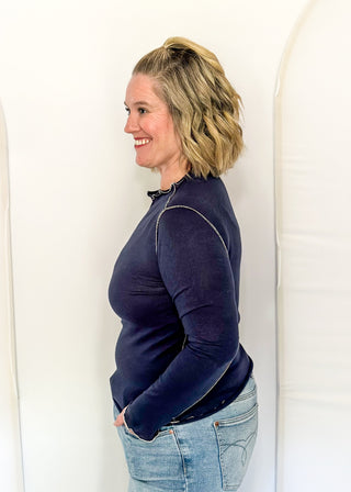 A woman with blonde hair is shown in profile wearing a dark navy long-sleeve top and light blue jeans, standing against a white studio backdrop with arched panels.