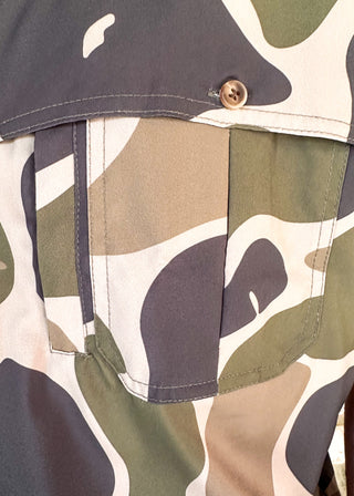 Close-up of a camouflage-patterned garment with a chest pocket and a single visible button closure. The fabric pattern mixes olive green, dark charcoal gray, tan, and off-white.