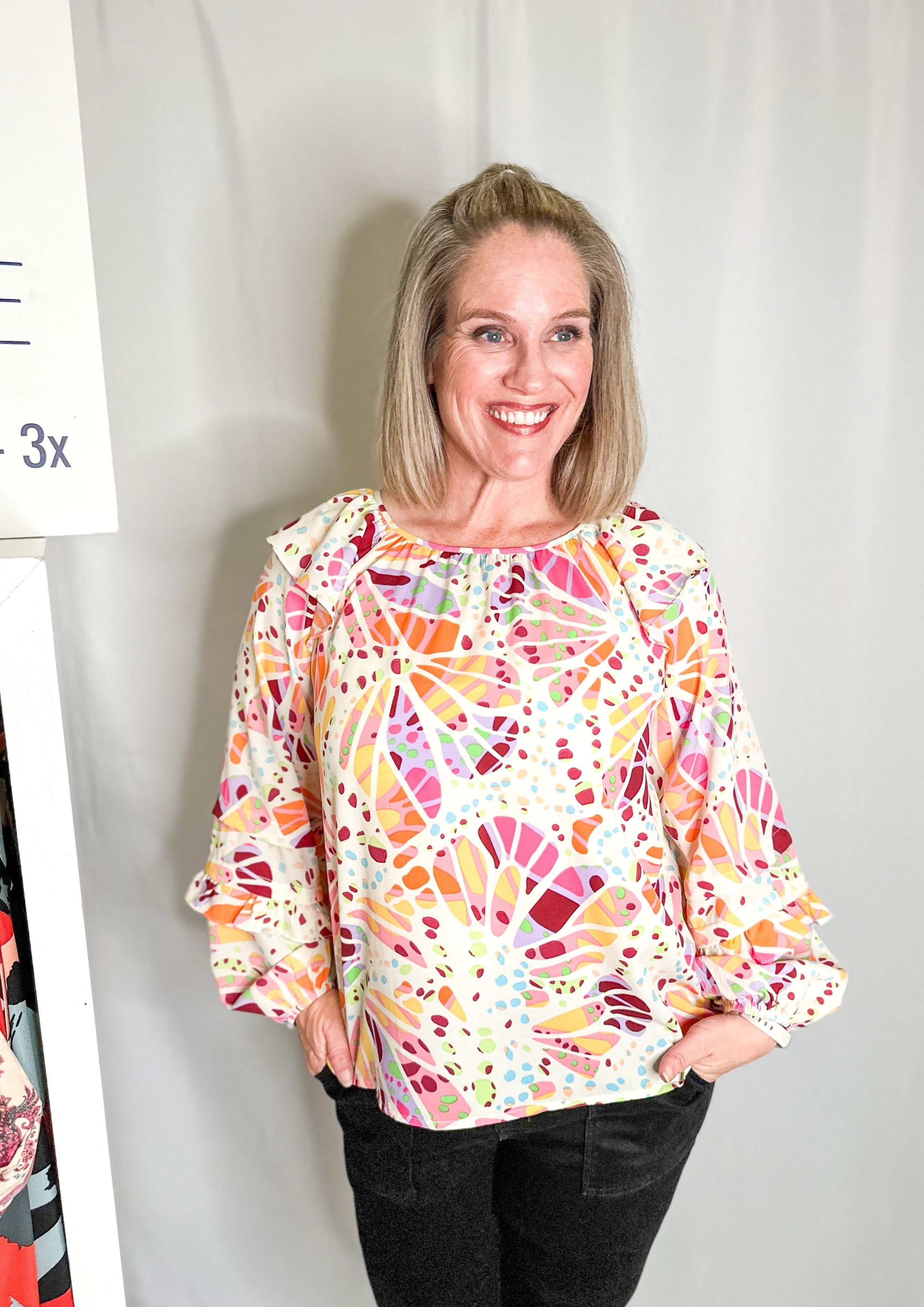 White long sleeve blouse with ruffle detail at the top of the sleeve near the shoulder and another small ruffle 3/4 down the sleeve. It has a higher neckline and multicolored abstract print all over.
