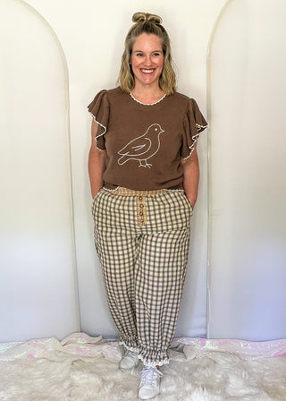 A woman stands indoors wearing a brown short-sleeve top with a white bird graphic and beige-and-white gingham pants, posed against a white arched backdrop.