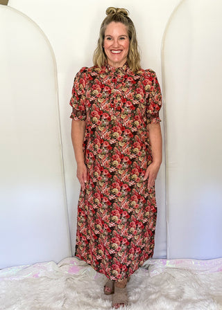 A woman stands in front of a white arched backdrop wearing a short-sleeve midi dress with a dense red, green, and beige floral print. She is standing on a white faux fur rug.