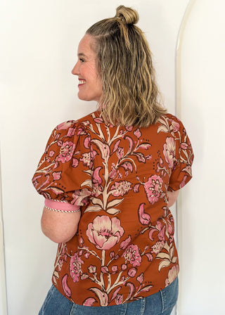 woman standing in a rust, pink and off white button up top and jeans.