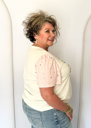 Off white short sleeve shirt with colorful embellished text and pink statement sleeves with decorative pearl studs.