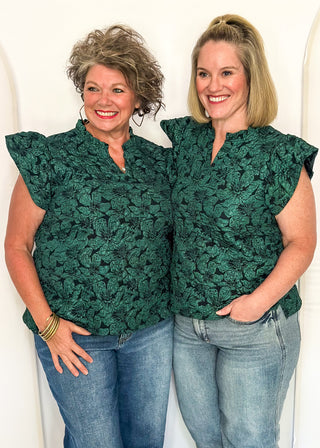 Two women wearing forest green jacquard flutter sleeve blouses with a floral pattern standing side by side