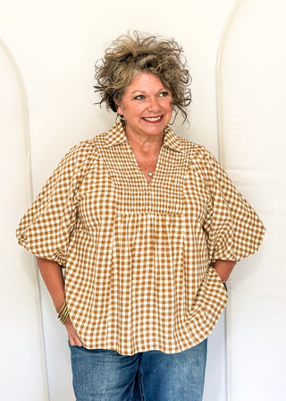 A smiling woman with short curly gray-blonde hair wearing a tan and white gingham puff-sleeve blouse and blue jeans, standing against a white arched backdrop.