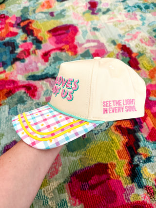 A hand holds a beige baseball cap with colorful text and a plaid brim, set against a vibrant, multicolored patterned textile background.